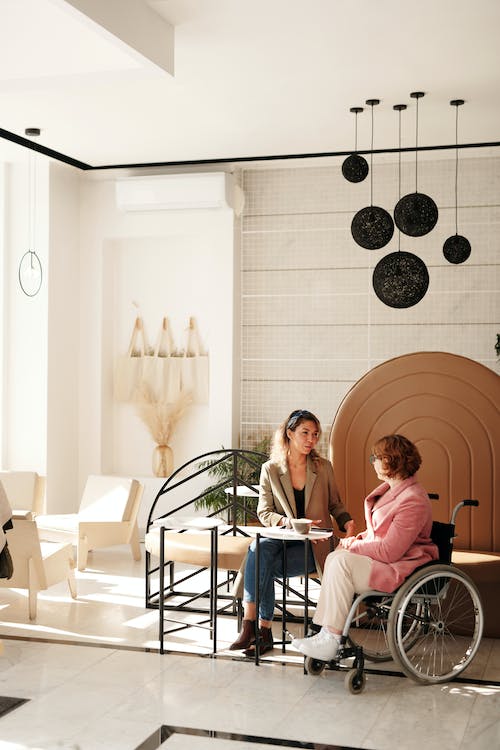 Dos mujeres sentadas tomando una bebida mientras hablan, una de ellas esta sentada sobre una silla de ruedas, la otra en una silla.